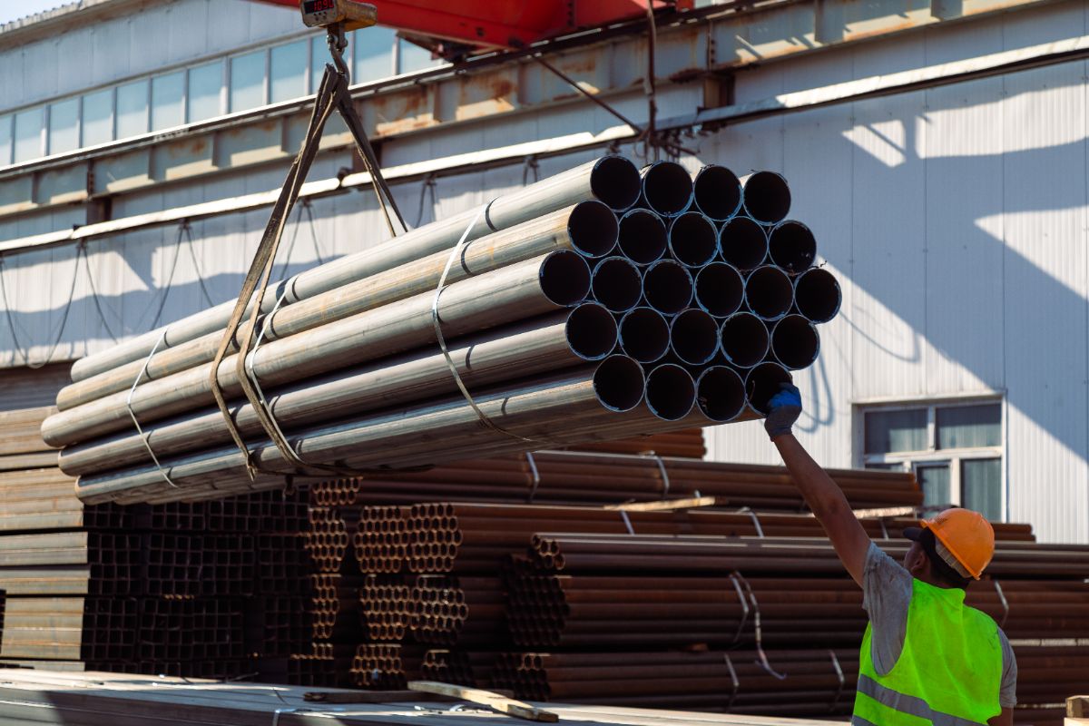 Worker guiding a lifted bundle of galvanized pipes at a steel supply yard