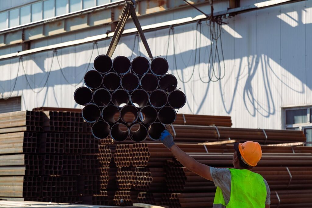 Worker guides a suspended bundle of steel pipes during warehouse handling and staging operations