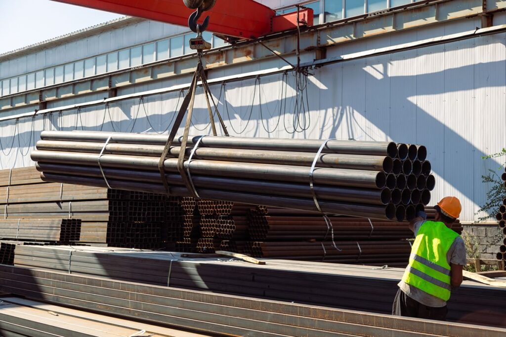 Worker guides a suspended bundle of steel pipes during material handling at an industrial facility.