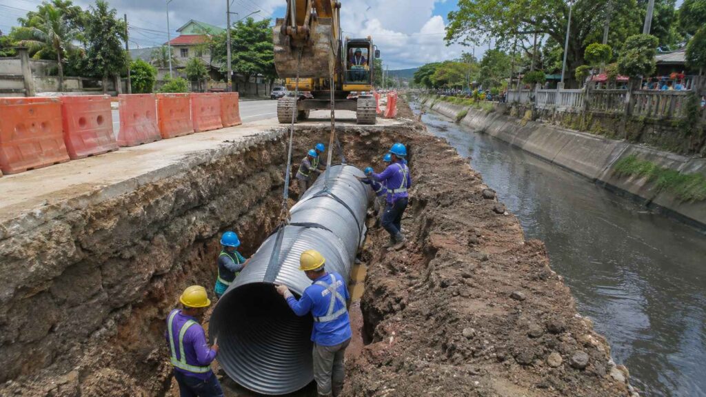 Construction workers install a large steel drainage pipe beside a roadside canal for flood and water control